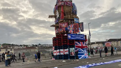 A large unlit bonfire covered in British and Northern Ireland flags beside a road. There are people milling around and cream-coloured blocks of flats in the background.