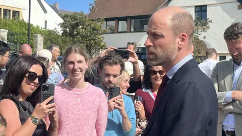 Prince William looking to the left as he talks to a crowd of people. There is a woman smiling in a pink jumper, and a woman with black hair and sun glasses taking a picture. 