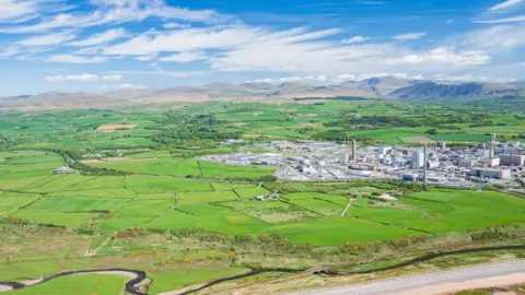 An aerial view of Sellafield and the space next to it, where the new Pioneer Park is planned. Sellafield looks like a very large industrial estate, with several buildings and cooling towers. Around it are green fields and the fells in the background.