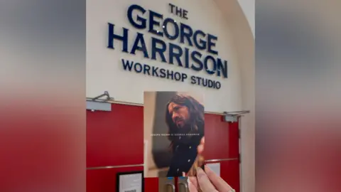 LIPA A postcard showing a film promo for the Sir Sam Mendes films about the Beatles showing George Harrison, held next to a sign for the George Harrison studio at LIPA 