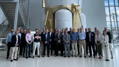 Space North East A delegation of space industry representatives standing in front of a Blue Moon lunar lander. The large craft is white with sections painted gold.