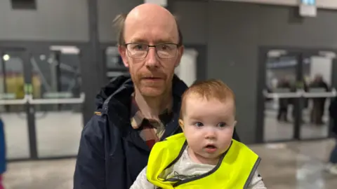 A bald man with square-rimmed glasses and a beard looks at the camera while holding a young baby who is wearing a grey shirt and yellow hi-vis jacket.