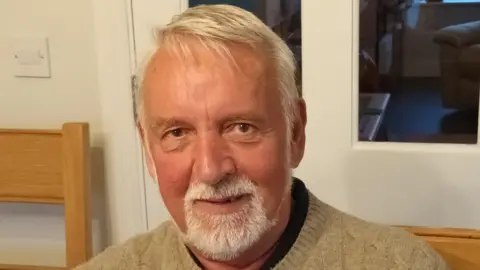 Brian Hughes A man with grey hair wearing a beige jumper smiles at the camera.