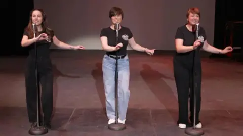 The Boogaloo Babes during rehearsals. The three women each stand behind a microphone stand while singing and dancing, their left arms outstretched.