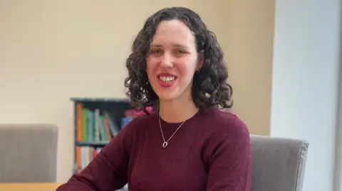 A woman with dark curly, shoulder-length hair looks into the camera. She is wearing a burgundy jumper with a silver necklace. She is sitting on a grey chair with a small bookcase in the background.