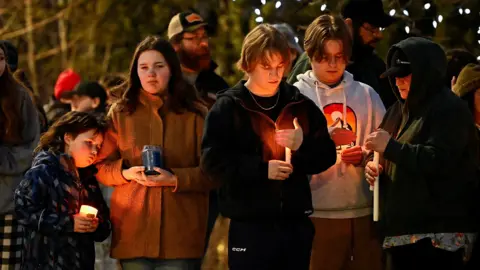 A row of five teenage mourners light candles near the scene. It is night time
