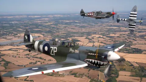 PA Media Three Spitfires fly in formation above the Kent countryside. The aircraft in the foreground bears shark-mouth nose art and the name "Grey Nurse", while two other Spitfires fly behind it over a patchwork landscape of fields, roads and villages.