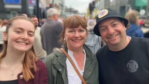 left to right: Eimear, Orla and John Penruddocke. Everyone is smiling looking directly into the camera, pictured outside with crowds of people in the background