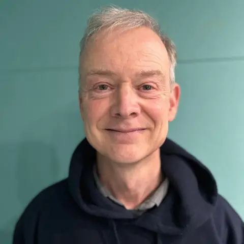 Grey-haired man smiling at the camera. He is wearing a blue hoodie with a grey shirt underneath. There is a green wall behind him.