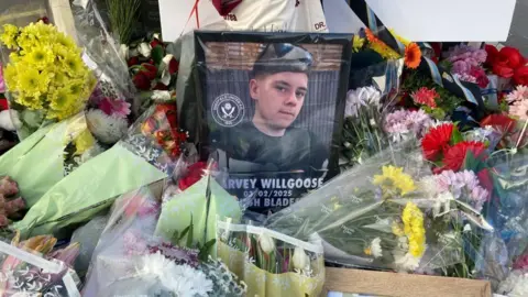 A framed photograph of a teenage boy with brown hair wearing a black T shirt and sunglasses. Around the frame are many bunches of flowers.