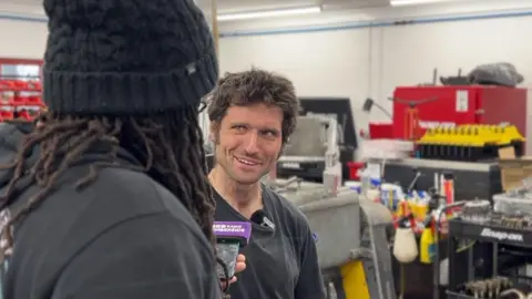 BBC A man with short dark hair and sideburns wearing a black T-shirt is talking into a phone with a purple microphone cover held by a man wearing a black woolly hat and with dreadlocked hair and wearing a black hoodie. They are standing in a workshop with heavy machinery in the background.