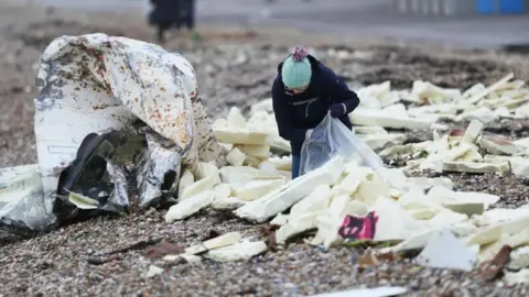 A person putting white blocks of foam into a large, clear plastic bag on a pebble beach