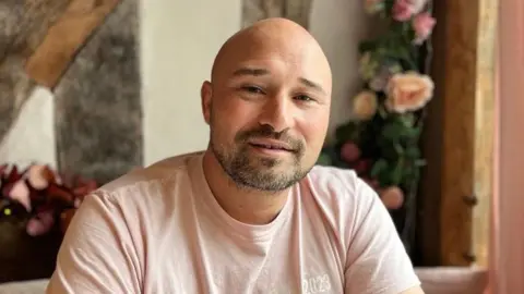 Jayson Moran, a 47-year-old bald man wearing a light pink T-shirt with branding for his bakery, called the The Crumbly Bun