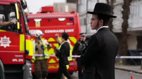 PA Media A man wearing a Jewish black hat and coat looks on as members of the London Fire Brigade respond to a blaze overnight