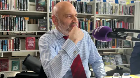 A man wearing a blue an white striped shirt sits in front of a bookshelf at a library. He is sitting in front of a purple microphone. He is holding his hand to his chin.