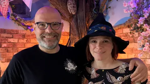 A man with a bald head, glasses and short beard is wearing a black t-shirt and smiling. He has his arm around a woman who is wearing a witch's hat and a black dress with an owl pattern. They are inside a cafe which is decorated with a fake tree and fake brick wall, bunting and fairy lights