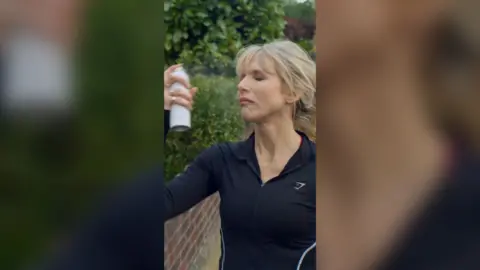 Lucy Punch as Amanda is wearing a black gym top and spraying a bottle of makeup setting spray on her face.
