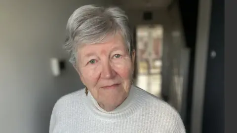 Sheila Jeffrey looking into the camera. She has short grey hair and is wearing a white jumper. 