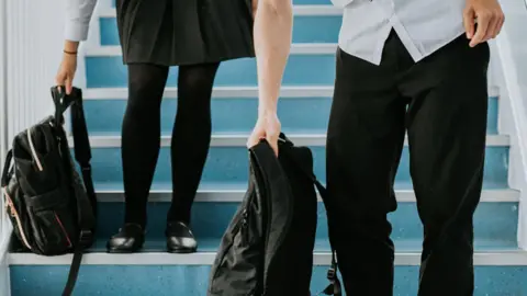 A close up stock image of two pupils lifting their backpacks from the blue stairs. The girl on the left is wearing a grey skirt, black tights and black flat shoes. The boy is wearing a short-sleeve white shirt, a pair of black trousers and a tie.