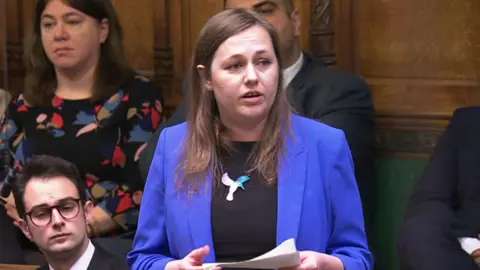 House of Commons A female MP stood up in the House of Commons speaking while holding a piece of paper