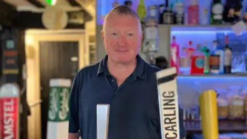 Jadzia Samuel/BBC Mark Cullinan, landlord of the Hare and Hound Pub in Godstone. Mark is stood behind the bar and is wearing a navy shirt.