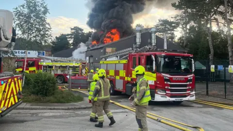 Hertfordshire Fire and Rescue Service A building on fire, with two fire engines outside it, with a large number of fire fighters walking past. Yellow pipes are on the ground. Flames are rising into the sky and trees are all around. 