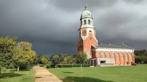 The Royal Victoria Hospital Chapel  - to the right of the shot - lit up by the sun but under dark, ominous clouds. There is a lawn in the foreground with a path leading to the chapel, with trees to the left of the frame and in front of the chapel, which is sideways on to the camera