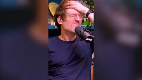 Comedian Elis James with his eyes closed and hands on his head. He is next to a BBC mic while recording a podcast