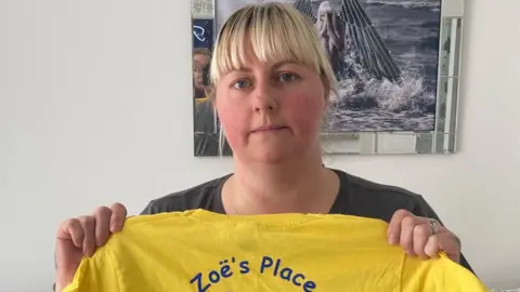 BBC Stephanie Perry, with blonde hair cut into a fringe, looks at the camera with a serious expression and holds up a yellow t-shirt with the Zoe's Place logo