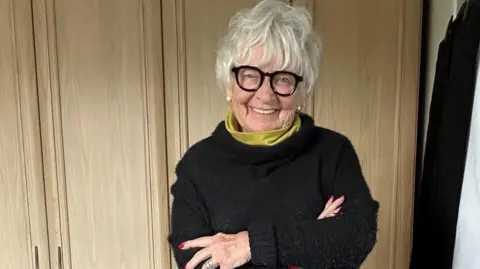 Vivian Truran Viv is pictured here posing for the camera. Her arms are crossed. She's wearing a black jumper with a lime green/yellow top underneath. She has short white hair and is wearing bold, black frames.