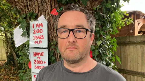 BBC/Sam Dixon-French Councillor John Trollope standing in front of the two oak trees in Willow Drive, Billingshurst. He is wearing a grey shirt and standing in front of demonstration placards, calling on the insurer Axa not to fell the trees.
