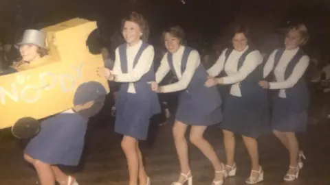 A grainy image, but in colour, showing five women in identical white tops and blue waistcoats who are marching in a conga line.