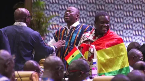 BBC An MP wrapped in a Ghanaian flag stands up in parliament - 22 October 2024