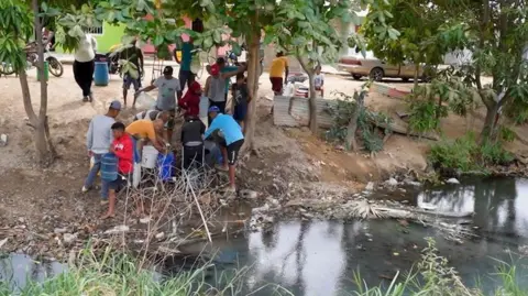 People fill plastic drums and containers with water from a stream. The water in the stream appears cloudy, and rubbish has collected at its banks.
