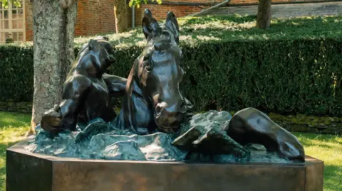 Longleat Safari Park Image of a large bronze sculpture of a horse and a man. In the background can be seen a clipped hedge and trees and a brick building.