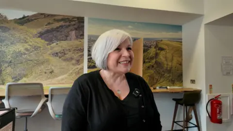 Julie Wright, a smiley woman with a white bob stands in front of the mock-up polling booth where staff are trained. She wears a black top and cardigan.