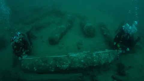 Underwater image of two divers measuring a barnacle-covered canon on the sea bed.