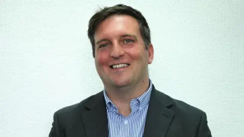 Stagecoach Group A head and shoulders picture of James Hanson, shown standing in front of a light green wall and smiling at the camera. He has short dark hair and is wearing a grey jacket and white and blue checked shirt. 