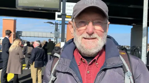 Robert Taylor is an older man with a white beard and moustache. He is wearing a blue cap and a blue jacket with a red shirt and is standing on the platform at Newsham station 