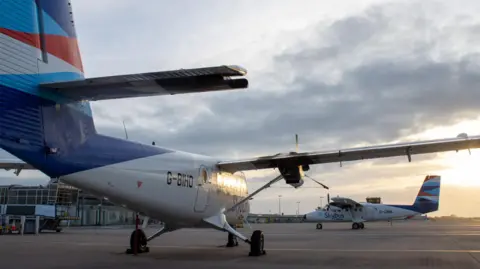 Two twin otter planes. One facing away to the camera and one facing diagonally. The two planes are on airport tarmac. The sky is grey and cloudy. 
