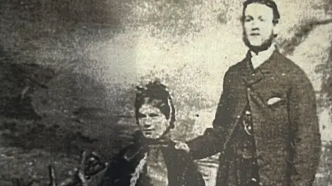 A faded black and white photograph shows a man and woman in Victorian attire.