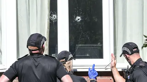 Three police officers in black uniforms and baseball style caps carry out a forensic examination. They are looking at two bullet holes in the window of a house 