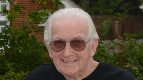 Ian Chatten smiles at the camera. He has short grey hair and wears sunglasses along with a black T-shirt. He is standing in front of a building with bushes and trees behind him.