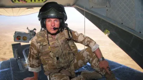 Antony Loveless Antony Loveless sitting in an army transport plane with the back hatch open. He is wearing brown camouflage gear and has a helmet on with a microphone to the front. At the back of the plane his camera is set up so he is taking photos of the ground below.