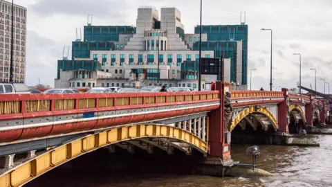 Getty Images Red and gold Vauxhall Bridge over the River Thames, packed with motor traffic, with the blocky, green-windowed MI6 building directly behind it.
