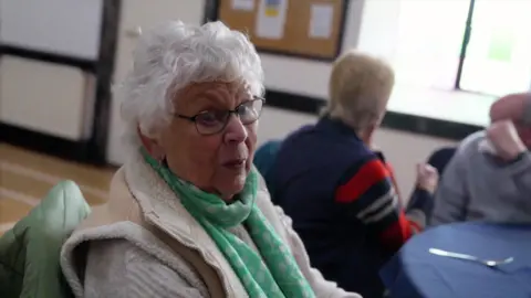 The image shows an elderly woman with white hair and glasses. She is seated at a community lunch. She is wearing a beige fleecy gilet, and a green and white dotted scarf