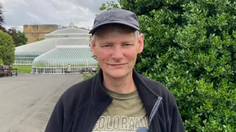 Allan Richardson - man in his 60s - with grey hair smiling at the camera. He is wearing a black baseball cap, a green t-shirt and black zip-up fleece. He is standing in front of the glass structure of the Kibble Palace in Glasgow's Botanic Gardens and there are trees and benches in the background.