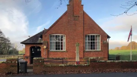 Scaleby Hall, a small red brick building, with the war memorial outside it.