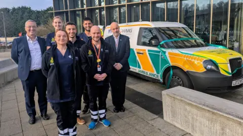 Students dressed in paramedic uniforms stand in front of an ambulance car. There are two women and five men pictured. Two of the men are in suits.