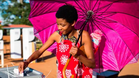 AFP/Getty Images A woman, holding a bright pink umbrella and wearing a red, patterned wrap around her body, places her vote into a ballot box. She has short hair and carries a baby on her back.